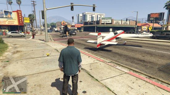 GTA 5 cheats - Franklin is standing next to a crossing. There is a bi-plane and a helicopter blocking traffic on the road. A market is on the other side of the road from Franklin.