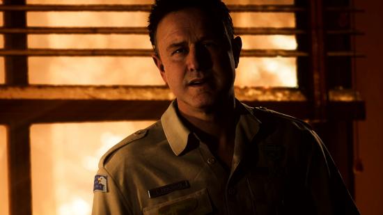 Chris Hackett, a character in The Quarry, wearing a law enforcement officer's uniform and is backlit by the light of a sunset coming through a weathered window behind him.