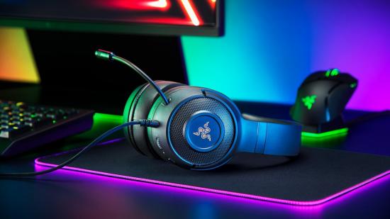 The Razer Kraken V3 X gaming headset sits atop a mouse mat, with a gaming keyboard, mouse, and monitor surrounding it