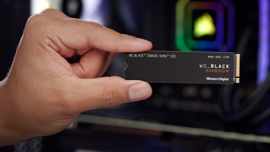 A hand (left) holds the WD Black SN850X NVMe SSD against a blurred background containing a gaming PC