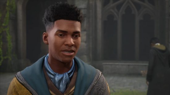 Hogwarts Legacy gameplay showcase: A Hogwarts studen wearing gold and blue-green robes stands in a grassy courtyard surrounded by grey gothic stone walls