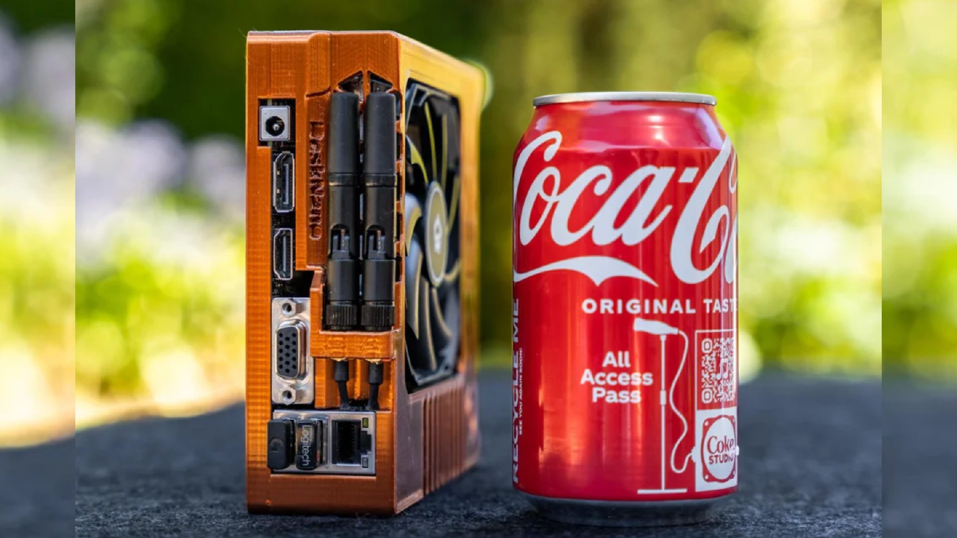 Diminuto PC para juegos con carcasa naranja junto a una lata de Coca-Cola en el jardín
