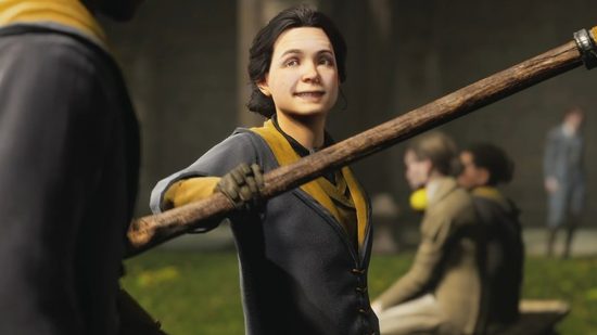 You can't pre-load Hogwarts Legacy on Steam or Epic, sorry: A white woman with dark hair wearing dark robes lined with gold holds out a broom with a smile to another person in a green field
