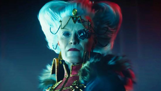 Warhammer 40k Darktide will get no new PC content until RPG gets fixed: An older woman with grey hair adorned with golden jewellery with a nose piercing and telescopic right eye looks into the camera wearing a fur lined red admiral jacket with gold trims