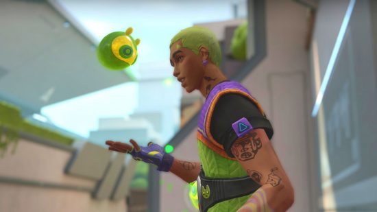 A Latino man with bright neon green hair wearing a neon green and purple tactical vest holds out his hand as a small green slime with a yellow superhero-style mask looks down at him