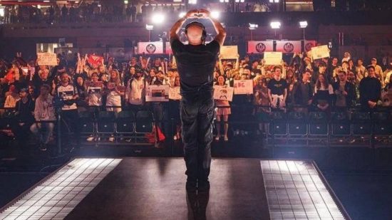 A bald man in front of a crowd wearing headphones forms a heart with his hands as they cheer and hold up signs