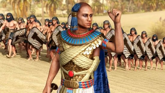 Total War Pharoah will not be owned by Microsoft, despite Sega talks: An Egyptian man in traditional gold and blue armor with blue braids and a shaven head holds up his fist as his army readies behind him in the sands