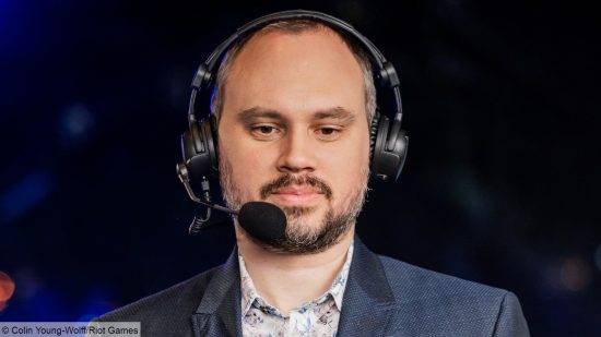 A bald man with a goatee wearing a suit and headphones stands looking down past the camera