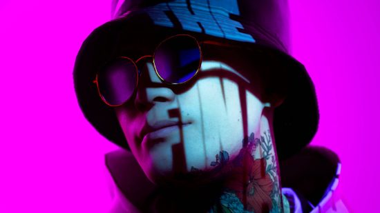 A man in sunglasses wearing a bucket hat with 'the finals' shining in light on his face smirks into the camera on a pink background