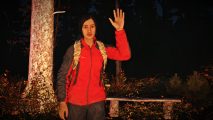 DayZ The Day Before: A woman with short black hair wearing a red and black coat with a camo backpack waves, a forest behind her
