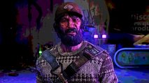 Dying Light 2 new game: A man with thick facial hair and a dirty red baseball cap looks ahead, a worn sweater and leather brown straps on his chest
