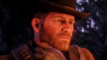 Red Dead Redemption 2 actor: a man with short brown stubble and a cowboy-like hat looks off to the side worriedly, as though he's thinking with a leather-gloved hand held up by his face