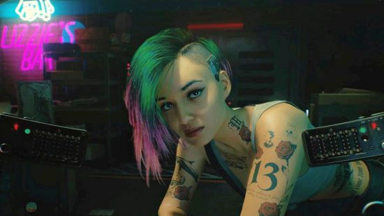 Cyberpunk 2077 2.1 patch notes: a young woman with green and pink hair leaning over a desk, arms crossed