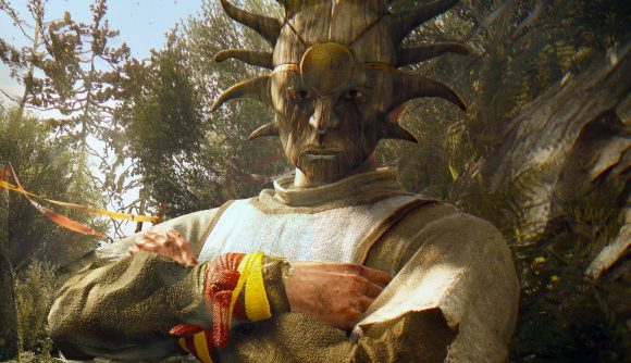 Dying Light has more players than its sequel, but dev says that's okay: A man wearing a wooden mask shaped like the sun stands in a forest location crossing his arms, colorful ribbons around his wrist