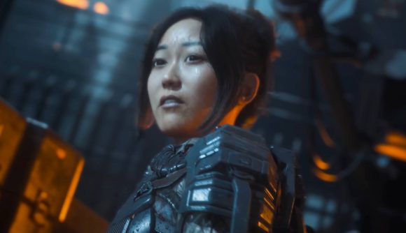 An Asian woman with her hair tied back in a bun wearing a combat space suit looks over her shoulder sweating