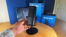 Elgato Wave:3 review image showing the mic on a desk near a human hand and its box.
