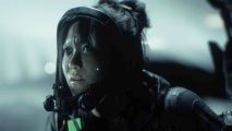 An Asian woman wearing a hazmat suit with her hair tied back looks upwards at something off camera