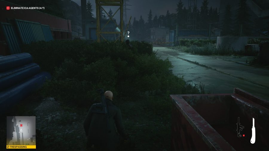 Outside, next to industrial buildings, Agent 47 watches a man concealed in foliage in Hitman 3