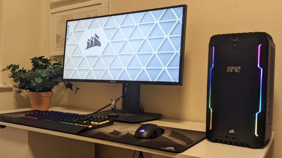 A Corsair One i300 gaming PC sits to the right of a gaming monitor, and other Corsair periperhals