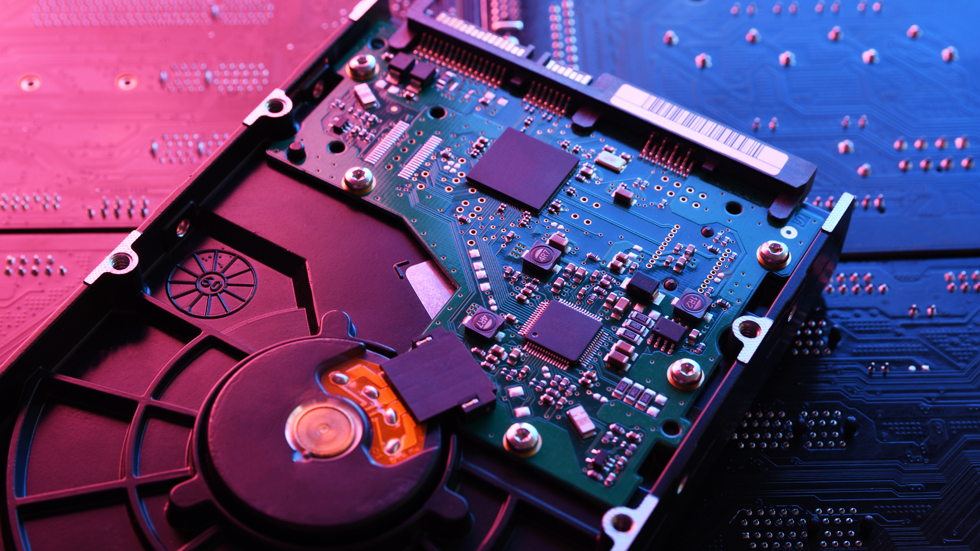 A mechanical hard drive lies on the desk face down, with blue and pink light shining on the HDD