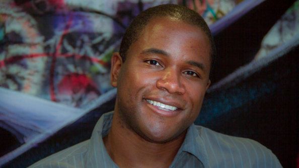 A headshot of Cardell Kerr, a young black man with a dreamy smile, broad nose, and hair shaved down to stubble. He is in front of a colorful background of what looks like concept art.