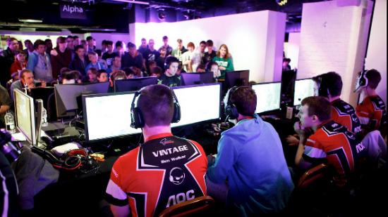 A group of competitive gamers in a small, crowded, brightly lit room, with their backs to the camera.