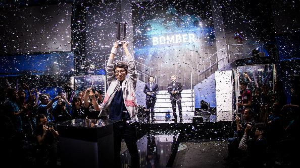 Bomber, a thin Korean pro player wearing a white zip-up team jacket, holds aloft the season 2 trophy with a look of confidence as confetti falls around him.